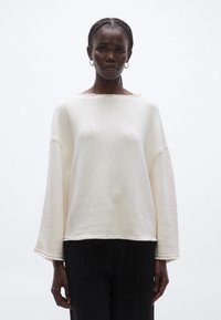 Krém színű, túlméretezett pulóver csónaknyakú kivágással, széles ujjakkal és nyers szegély részletekkel, puha anyagból készült. Fekete nadrággal párosítva.