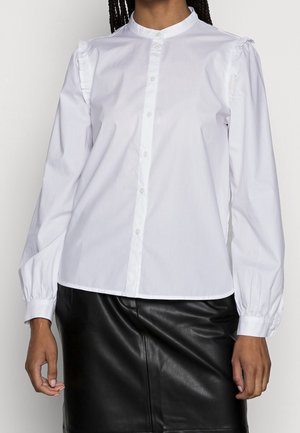 Woman wearing a white long-sleeve button-up blouse with ruffled shoulder detail and a black leather skirt.