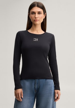 Vrouw met stijlbruin haar, gekleed in een zwarte top met lange mouwen met een klein wit logo op de borst en blauwe spijkerbroek, staand tegen een effen achtergrond.