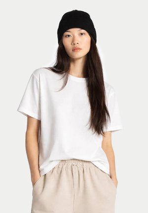 Junge Frau trägt eine schwarze Strickmütze, weißes kurzärmeliges T-Shirt und beige Hose, mit Händen in den Taschen, vor einfachem Hintergrund.