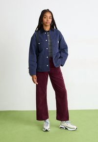 Giovane donna con lunghe trecce indossa una giacca blu scuro con bottoni, una camicia a righe, pantaloni in velluto a coste bordeaux e sneakers bianche, in piedi su un pavimento verde contro un muro bianco.