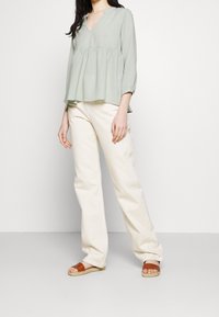 Donna che indossa una blusa larga verde chiaro, pantaloni a gamba larga bianco sporco e sandali marroni con le punte scoperte, in piedi contro uno sfondo bianco semplice.