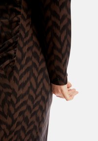 Braunes Kleid mit Zickzackmuster, seitlichem Raffen, langen Ärmeln und einem Knopfakzent am Bündchen. Glatte Textur.