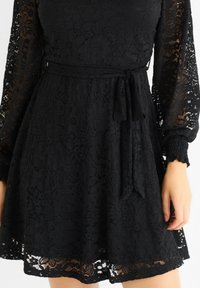 Black lace dress with long, sheer sleeves, floral pattern, a belted waist, and a scalloped hemline. Textured fabric with a slight sheen.