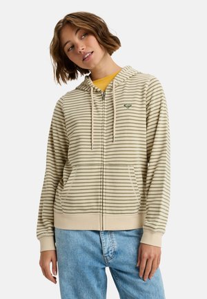Haut à capuche beige zippé avec des rayures horizontales vertes, capuche à cordon, poches kangourou et un petit logo sur la poitrine. Porté par-dessus une chemise jaune.