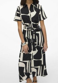 Vestido de camisa de manga corta en negro y crema con patrones geométricos, con botones en el frente, cintura ceñida y aberturas laterales. Textura de tela suave.