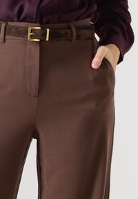 Bruine op maat gemaakte broek met een gladde textuur, glad voorkant en zijzakken, gecombineerd met een donkerbruine suède riem met een gouden gesp.