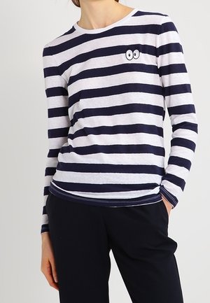 Chemise à manches longues blanche avec des rayures horizontales bleu marine, arborant un petit design brodé d'yeux sur la poitrine. Tissu en coton doux.