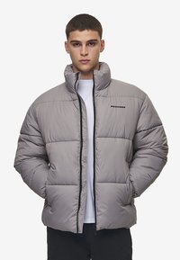 Grå quilted jakke med høj krave, lynlås og syede paneler. Mærket "PEGAADOR" synligt på brystet. Glat tekstur.
