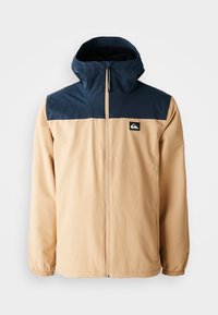 Beige jacket with navy blue shoulder and hood area, full front zipper, elastic cuffs, and a small logo patch on the left chest.