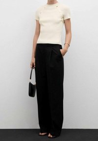 Žena v krémovém krátkém rukávu s žebrovaným topem a knoflíky, v širokých černých kalhotách s vysokým pasem, držící černou kabelku.