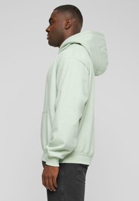 Mintgrønn hettegenser laget av myk stoff, med en romslig hette, frontlomme og ribbede mansjetter og kant. Bære med mørke jeans.