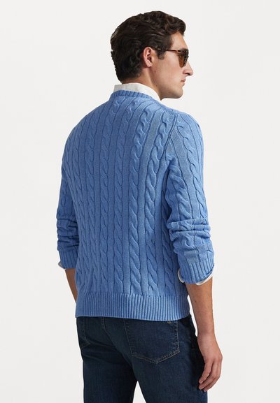 Blauwe kabelgebreide trui met geribde manchetten en zoom. De achterkant heeft een gestructureerd patroon. Draag over een witte blouse met kraag.