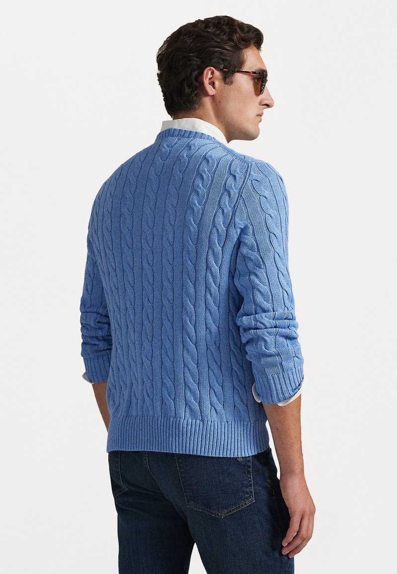 Pull en maille bleue avec des poignets et un bas côtelés. Le dos présente un motif texturé. Porté par-dessus une chemise blanche à col.