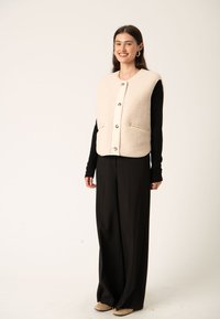 Beige uldvest med rund hals, frontknapper og to sidelommer, båret over en sort langærmet bluse og parret med sorte bukser.