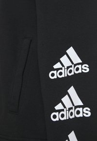 Felpa nera con cappuccio realizzata in un tessuto liscio, caratterizzata da loghi Adidas bianchi in un motivo geometrico ripetuto lungo la manica e una tasca laterale.