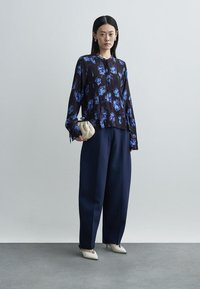 Schwarze Bluse mit Blumenmuster und blauen Akzenten, lange Ärmel, kombiniert mit navyblauen, weit geschnittenen Hosen. Hält eine cremefarbene Clutch-Tasche, trägt weiße spitze Schuhe.