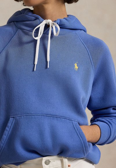 Mėlynas džemperis su gobtuvu, baltos virvelės, ant krūtinės geltonai siuvinėtas logotipas, vilkimas su baltais kelnėmis, ranka kišenėje priekyje.