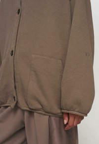 Braune, schicke Jacke aus weichem Stoff, mit einer aufgesetzten Tasche, schwarzen Knöpfen und gerafften Manschetten für einen entspannten Sitz.