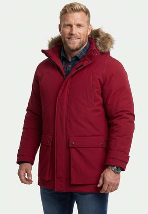 Mann trägt roten Wintermantel mit Fellkapuze und blau kariertes Hemd, lächelnd, steht mit entspannten Händen neben dem Körper vor weißem Hintergrund.