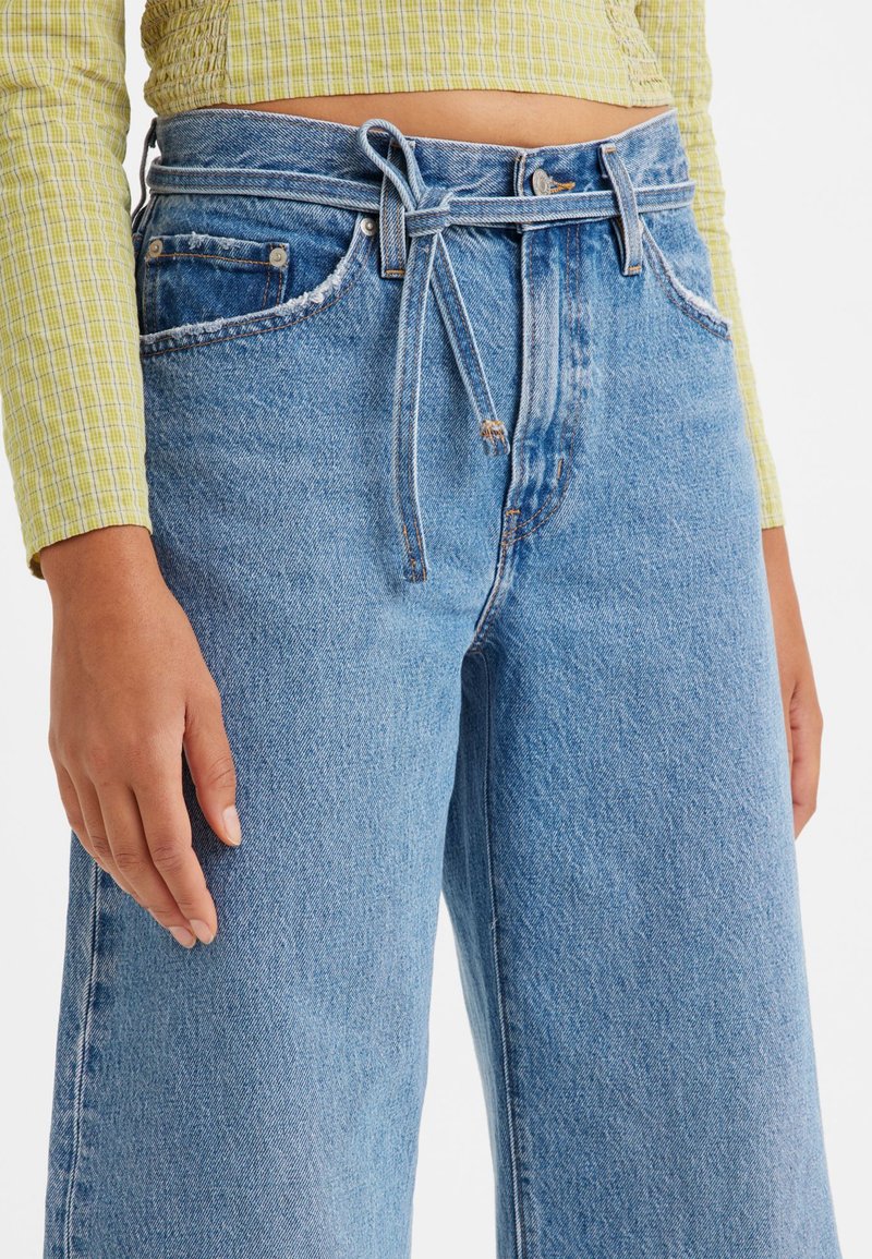 Hellblaue High-Waist-Weitbeinhosen aus Denim mit einem taillierten Bund, ausgefransten Taschenrändern und einer glatten Textur.