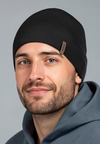 Zwarte gebreide muts met een aansluitende pasvorm, gemaakt van zacht stof. Voorzien van een klein bruin label aan de zijkant. Draagt een persoon met een grijze hoodie.