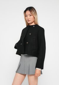 Femme aux longs cheveux portant une veste cropped noire à double boutonnage sur une jupe plissée gris clair, sur fond blanc uni.