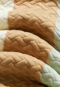 Tissu tricoté doux avec des motifs en zigzag et géométriques en relief dans des tons beige, crème et vert clair, montrant des plis et la texture.