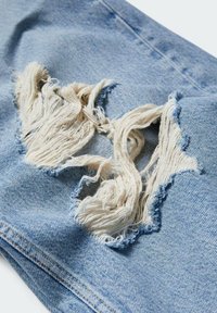 Tissu en denim bleu clair avec un grand trou effiloché révélant des fils lâches, surface texturée et zones légèrement décolorées.
