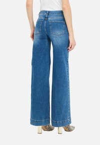 Jeans de denim azul de pierna ancha con cintura alta, con bolsillos traseros y dobladillo sin terminar. Combinados con botas de tobillo con estampado.