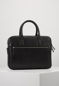 Serviette en cuir noir avec deux poignées et une poche zippée horizontale à l'avant, posée sur une surface blanche contre un fond uni.