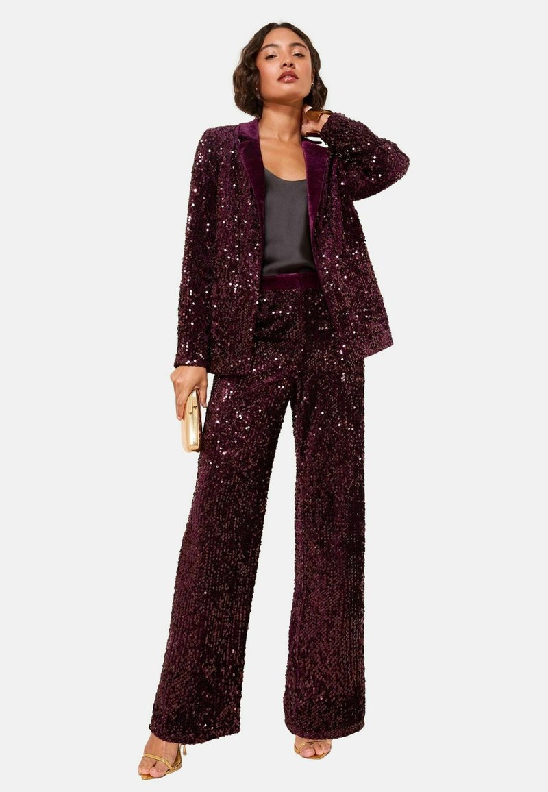 Sequin-embellished suit in deep purple with a fitted blazer and wide-leg pants. Features a contrasting dark satin inner collar. Gold clutch accessory.