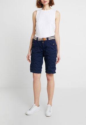 Femme portant un haut blanc sans manches, un short bleu marine longueur genoux avec une ceinture rayée, et des baskets blanches, debout devant un fond uni.