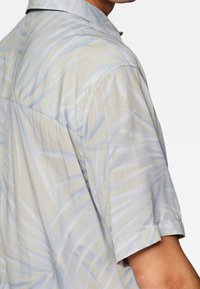 Camisa de manga corta azul claro con un sutil patrón de hojas de palma, usada por una persona que muestra el hombro y la parte superior del brazo contra un fondo blanco.