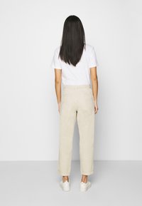 Pantaloni beige, larghi e dalla consistenza liscia, con orli cuffati e due tasche posteriori. Indossati con una maglietta bianca semplice e sneakers bianche.