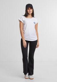 Hvid kortærmet t-shirt med optrukne ærmer, prydet med et blåt logo. Kombineret med sorte flagrende bukser og hvide sneakers. Sort kasket båret.