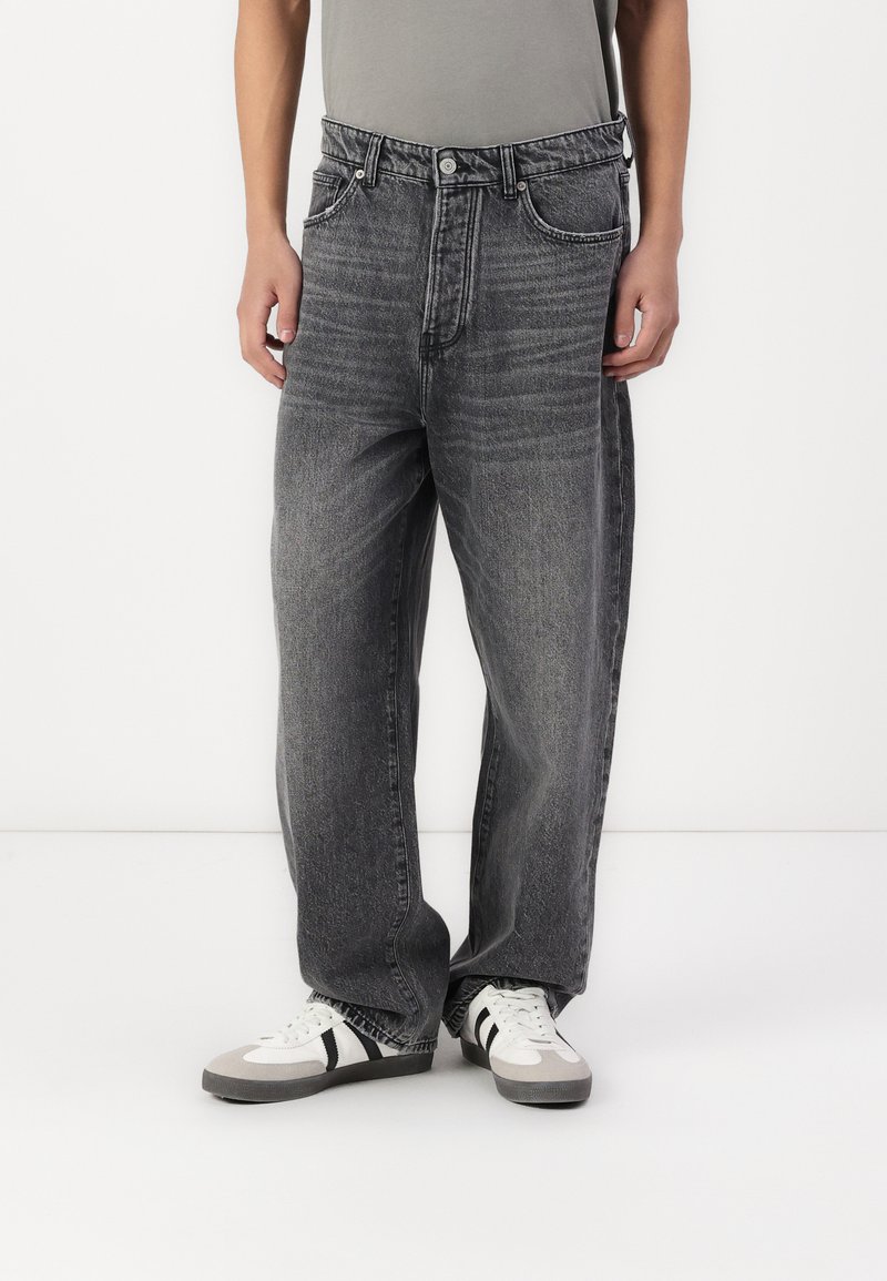 Baggy black denim jeans with a faded finish. Distressed texture, five-pocket design, and a button closure. Worn with gray and white sneakers.