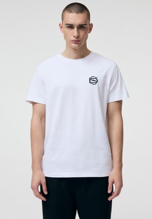 Młody mężczyzna z krótkimi włosami nosi gładką białą koszulkę z krótkim rękawem, z czarnym stylizowanym logo "S" na lewej piersi, w połączeniu z ciemnymi spodniami.