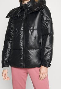 Černá prošívaná bunda s lesklým povrchem, vysokým límcem, zipovým zapínáním a knoflíkovými detaily na oversized límci a manžetách.