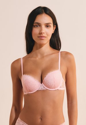 Mujer con cabello largo y oscuro que lleva un sujetador de encaje rosa claro y ropa interior a juego sobre un fondo beige liso.