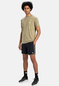 Hombre de pie con la mano en el bolsillo, vistiendo una camiseta gráfica verde oliva, pantalones cortos deportivos negros con ribete, calcetines negros y zapatillas de correr negras.