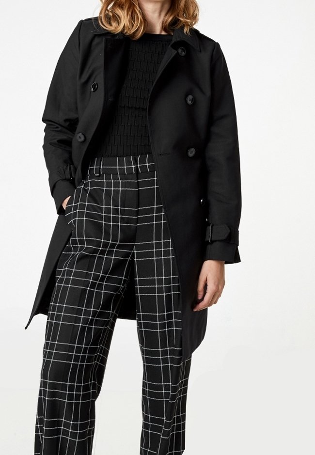 Femme portant un manteau noir croisé, un pull texturé noir et un pantalon noir à motif carreaux blanc, debout, une main dans la poche.