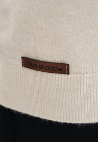 Kremowy sweter dzianinowy z prążkowanym wykończeniem, z brązową skórzaną metką z napisem "blue shadow" w dolnej części.