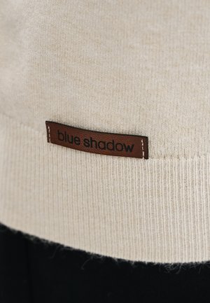 Beżowa dzianina z brązową skórzaną metką wszytą, z wytłoczonym czarnym napisem "blue shadow."