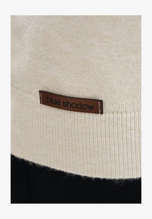 Beżowa dzianina z brązową skórzaną metką wszytą, z wytłoczonym czarnym napisem "blue shadow."