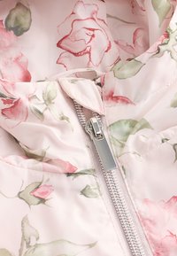 Tissu à motif floral en rose pâle avec des feuilles vertes. Dispose d'une fermeture éclair argentée sur le devant avec un col montant et une texture légère.