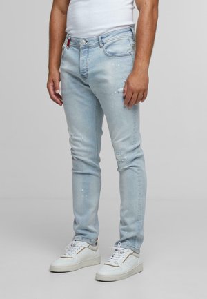 Light blue denim jeans with distressed details and subtle paint splatters. Fitted design with five pockets and a button fly. Paired with white sneakers.