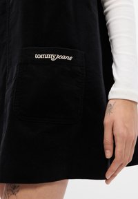 Μαύρο φόρεμα από κοτλέ με μπροστινή τσέπη που έχει το κεντημένο λογότυπο "tommy jeans". Το φόρεμα έχει ευθεία γραμμή και υφή από καπιτονέ ύφασμα.