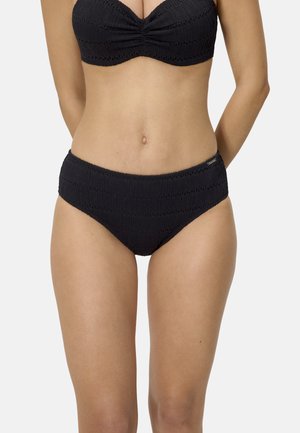 Mujer con bikini negro texturizado, con parte inferior y superior a juego, de pie con los brazos detrás de la espalda sobre un fondo claro y liso.