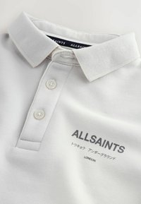 Bílé polo tričko AllSaints se dvěma knoflíky a černým logem s textem včetně japonských znaků a nápisem „LONDON“ na hrudi.